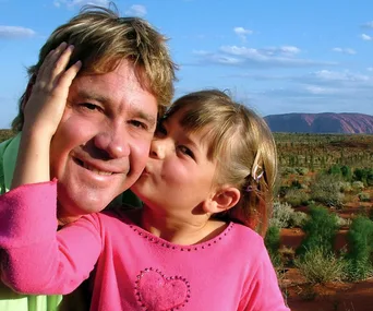 Father and young daughter share a sweet moment in a scenic outdoor setting with blue sky and landscape in the background.