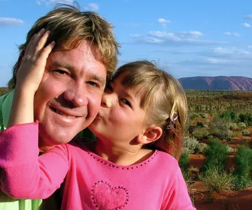 Father and young daughter share a sweet moment in a scenic outdoor setting with blue sky and landscape in the background.