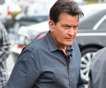 Man in a blue shirt walking outdoors, with a serious expression, cars in the background.