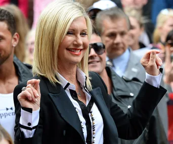 Olivia Newton-John smiling in a crowd, wearing a black blazer and white shirt.