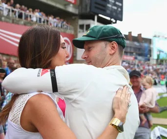 Man in cricket attire embracing a woman at a stadium with a crowd in the background.