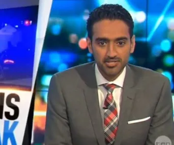 News anchor presenting on set with a blurred background, wearing a checkered tie and gray suit.