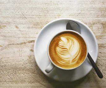 A cup of coffee with latte art, featuring a leaf pattern, on a saucer with a spoon on a wooden table.