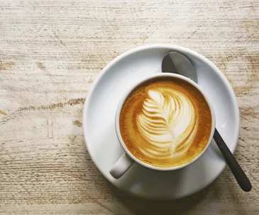 A cup of coffee with latte art, featuring a leaf pattern, on a saucer with a spoon on a wooden table.