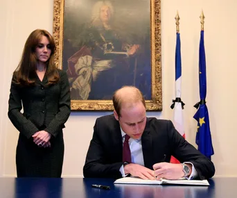 Prince writing in condolence book, woman stands beside; French and EU flags behind in solemn setting.