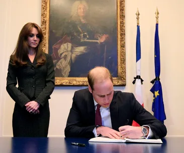 Prince writing in condolence book, woman stands beside; French and EU flags behind in solemn setting.