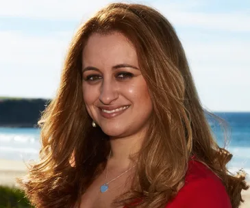A woman smiling at the camera by the beach, wearing a red top and pendant necklace.