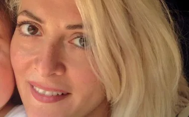 Blonde woman smiling with light-colored hair and brown eyes, partially cropped, against a dark background.