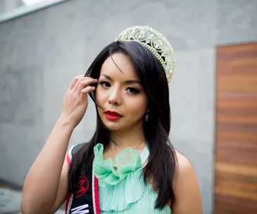 Beauty queen wearing a crown and sash, adjusting her hair, standing against a plain background.