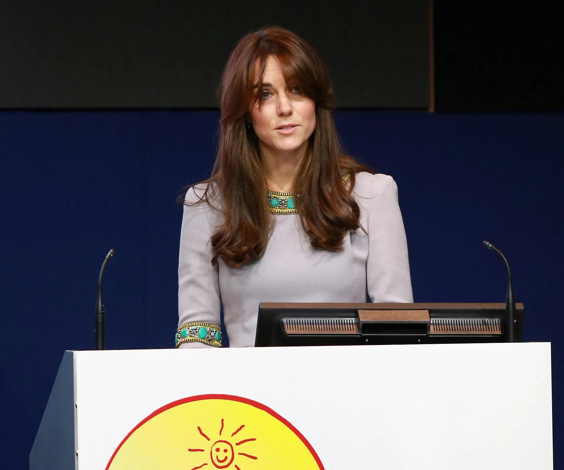A woman with long hair stands at a podium with a smiling sun emblem, speaking at an event.