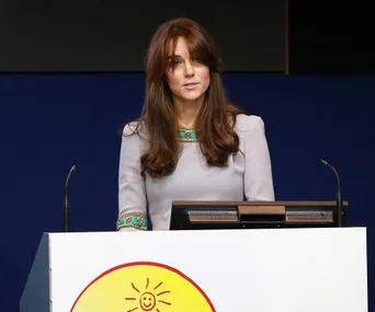 A woman with long hair stands at a podium with a smiling sun emblem, speaking at an event.