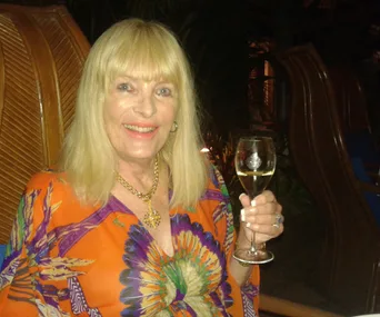 A smiling woman with long blonde hair holds a glass of wine, wearing a vibrant, colorful dress and gold necklace.