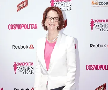 A woman in a white blazer and pink top smiles at the Cosmopolitan Women of the Year event backdrop.