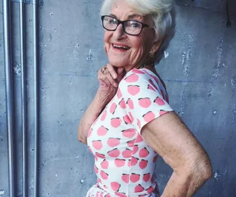 Elderly woman with glasses, smiling, wearing a peach-patterned dress against a concrete background.