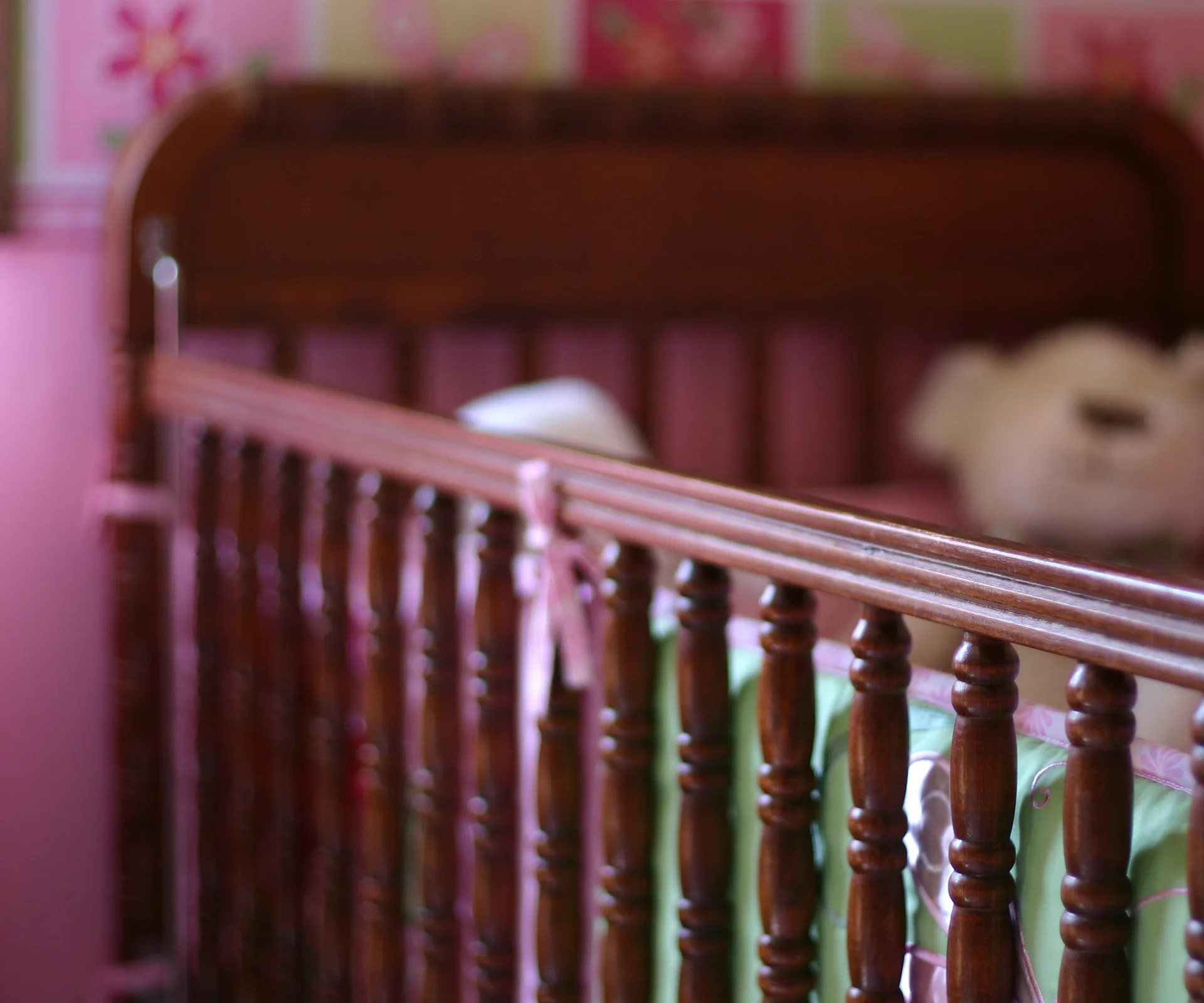 A wooden crib with pink bedding and a teddy bear in a softly lit pink nursery.