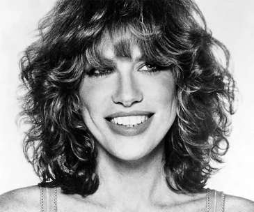 Carly Simon smiling in a black and white portrait with curly hair.