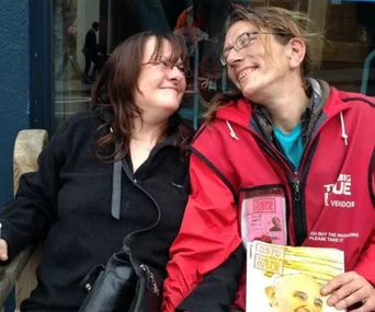 A man in a red Big Issue jacket smiles at a woman; he's holding a Big Issue magazine, both appear to be happy.
