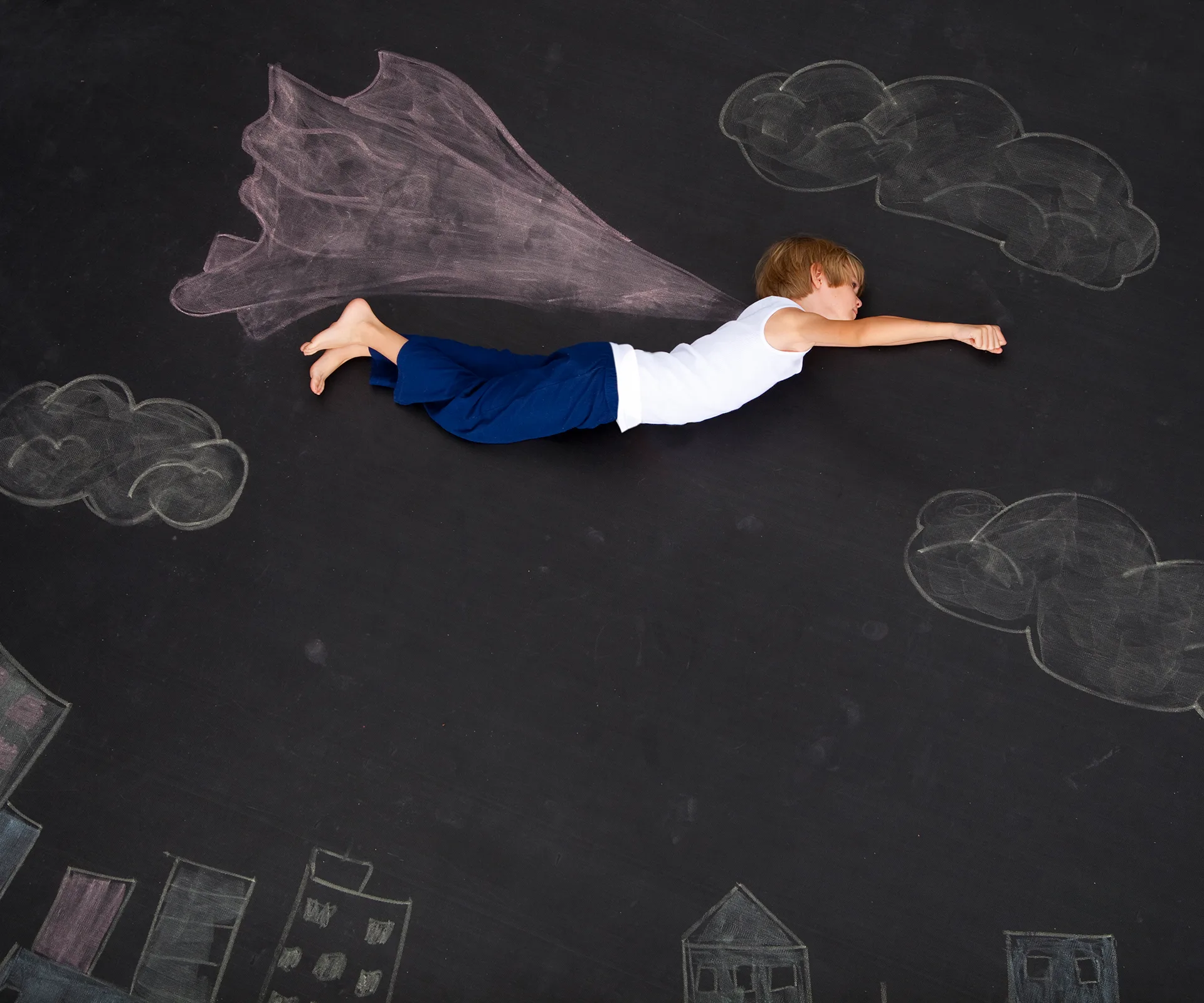 A young boy pretends to fly like a superhero, lying on a chalk drawing of a cityscape with clouds and a cape.
