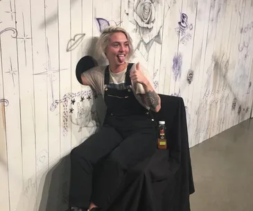 Person with arm in wall hole for tattoo, smiling, thumbs up, wearing overalls, surrounded by sketches.