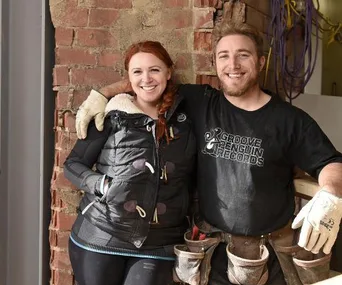 Two people smiling, wearing work gear, leaning against a brick wall in a renovation setting.