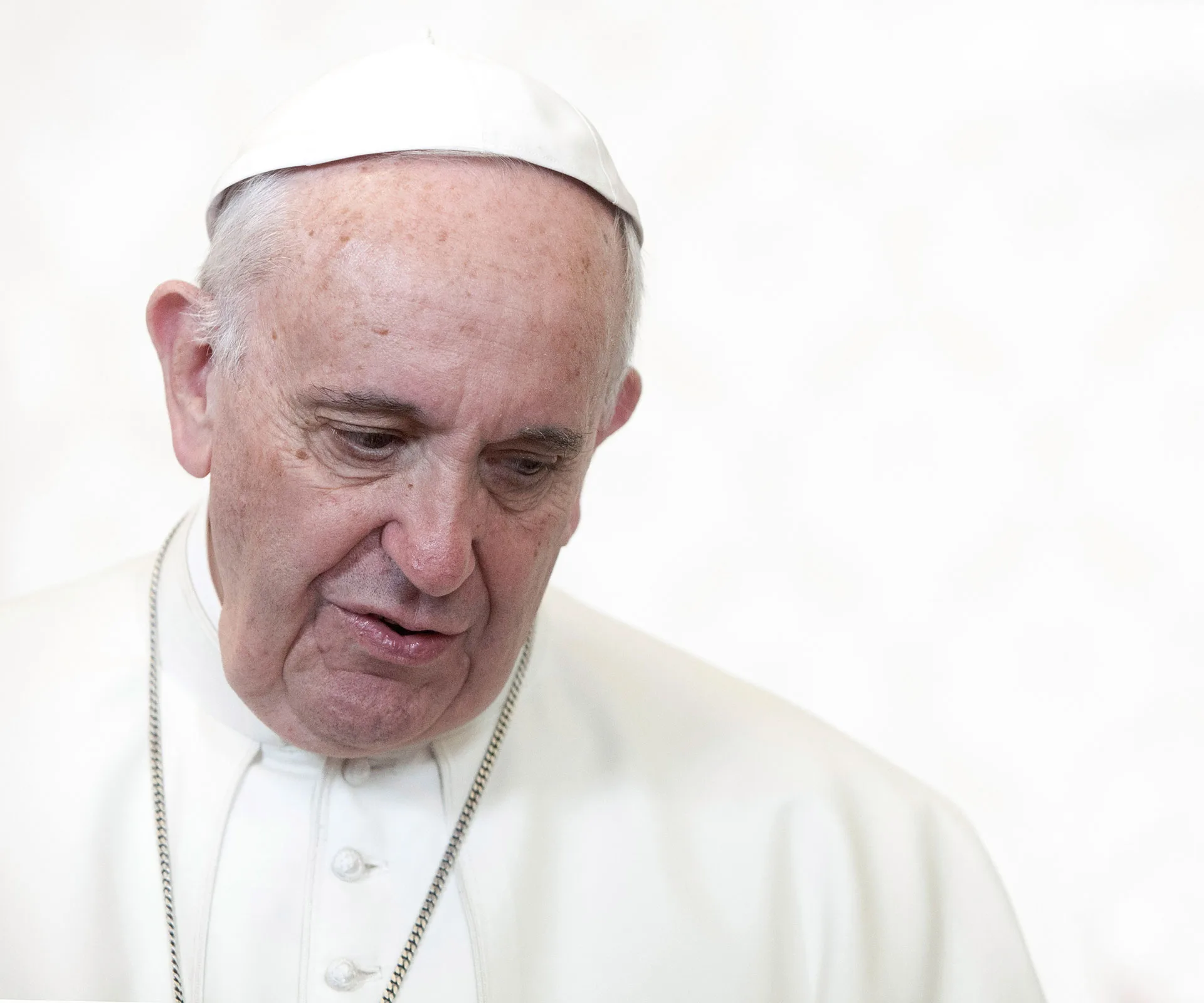 Pope wearing a white cassock and cap, looking downward against a light background.