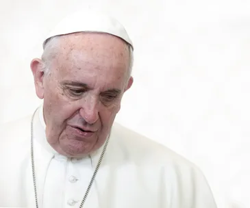 Pope wearing a white cassock and cap, looking downward against a light background.
