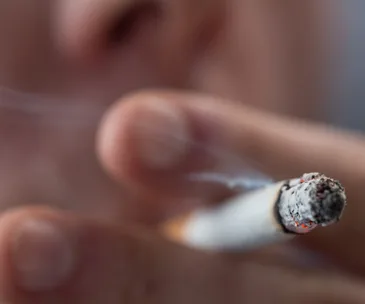 Close-up of a person holding a lit cigarette, with smoke visible and the face blurred in the background.