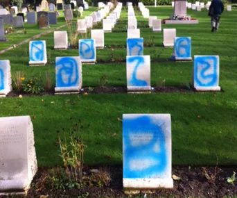 Headstones in a cemetery vandalized with blue spray paint graffiti.