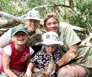 Family in outdoor setting, wearing hats, holding a small crocodile, with trees in the background.
