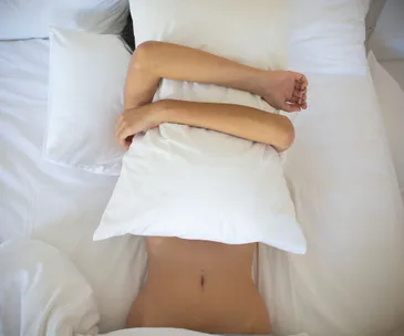 Person lying in bed covering face with a pillow, arms wrapped around it, in a brightly lit room.