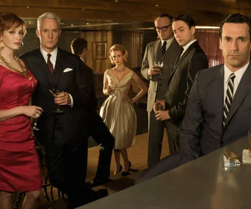 A group of six well-dressed individuals in 1960s-style attire at a dimly lit bar, some holding drinks.