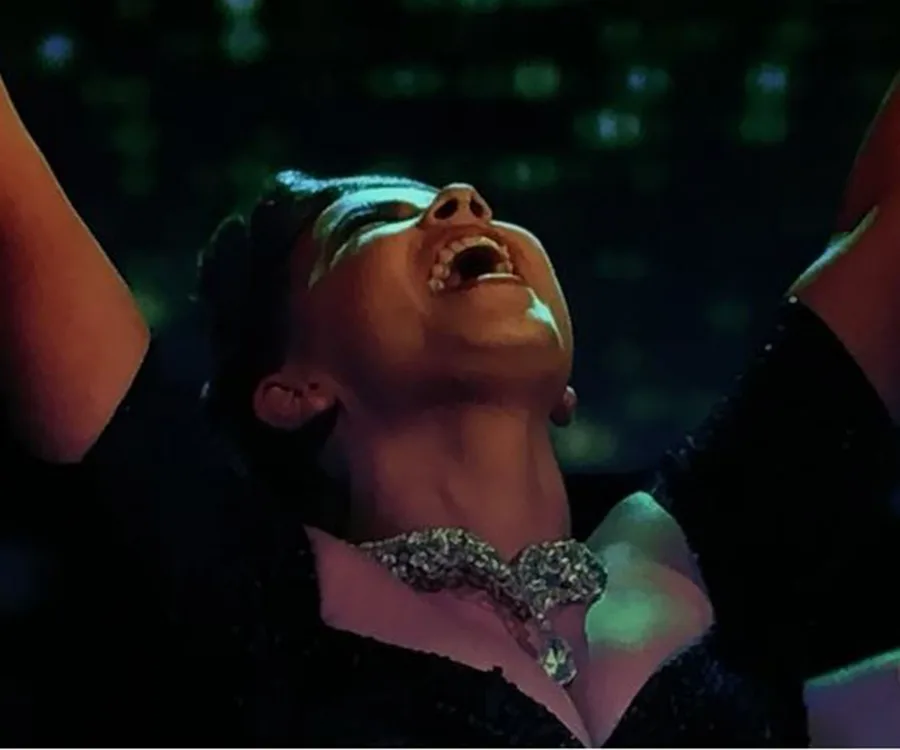 Dancer celebrates victory, head tilted back, arms raised, joy on face, wearing a sparkling necklace and dark dress.