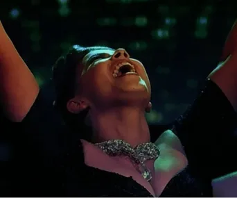 Dancer celebrates victory, head tilted back, arms raised, joy on face, wearing a sparkling necklace and dark dress.