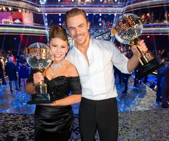 Two "Dancing with the Stars" winners celebrate on stage holding their trophies, surrounded by confetti and audience.