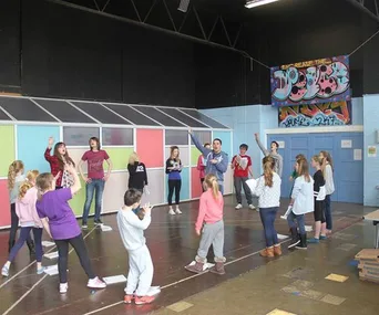A group of children and adults participating in a theater or dance rehearsal in a school gym with colorful backdrops.