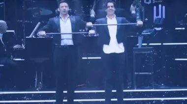 Two men in suits on stage perform a comedic act with feather dusters in the background.