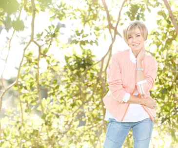 A smiling woman in a peach jacket and jeans stands outdoors, surrounded by green leaves and bright sunlight.