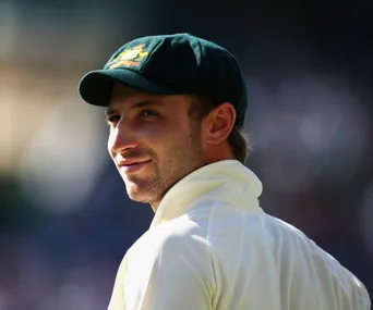 "Cricketer in white uniform and cap looking back, under bright sunlight."