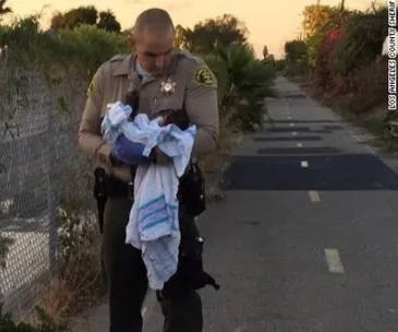 Sheriff's deputy holding a swaddled baby on a bike path, with greenery and a fence in the background.
