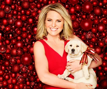 Woman in a red dress holding a puppy with a red ribbon, against a backdrop of red Christmas ornaments.