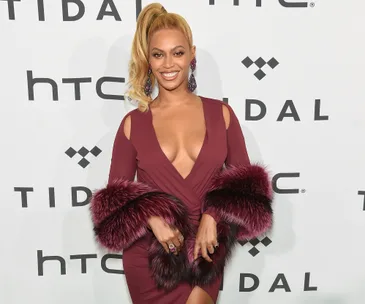 "Person in a burgundy dress with fur accents, smiling at a Tidal event backdrop."