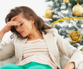 Woman in beige blazer appearing stressed, sitting with hand on forehead, a holiday-decorated tree in the background.