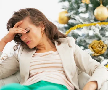 Woman in beige blazer appearing stressed, sitting with hand on forehead, a holiday-decorated tree in the background.