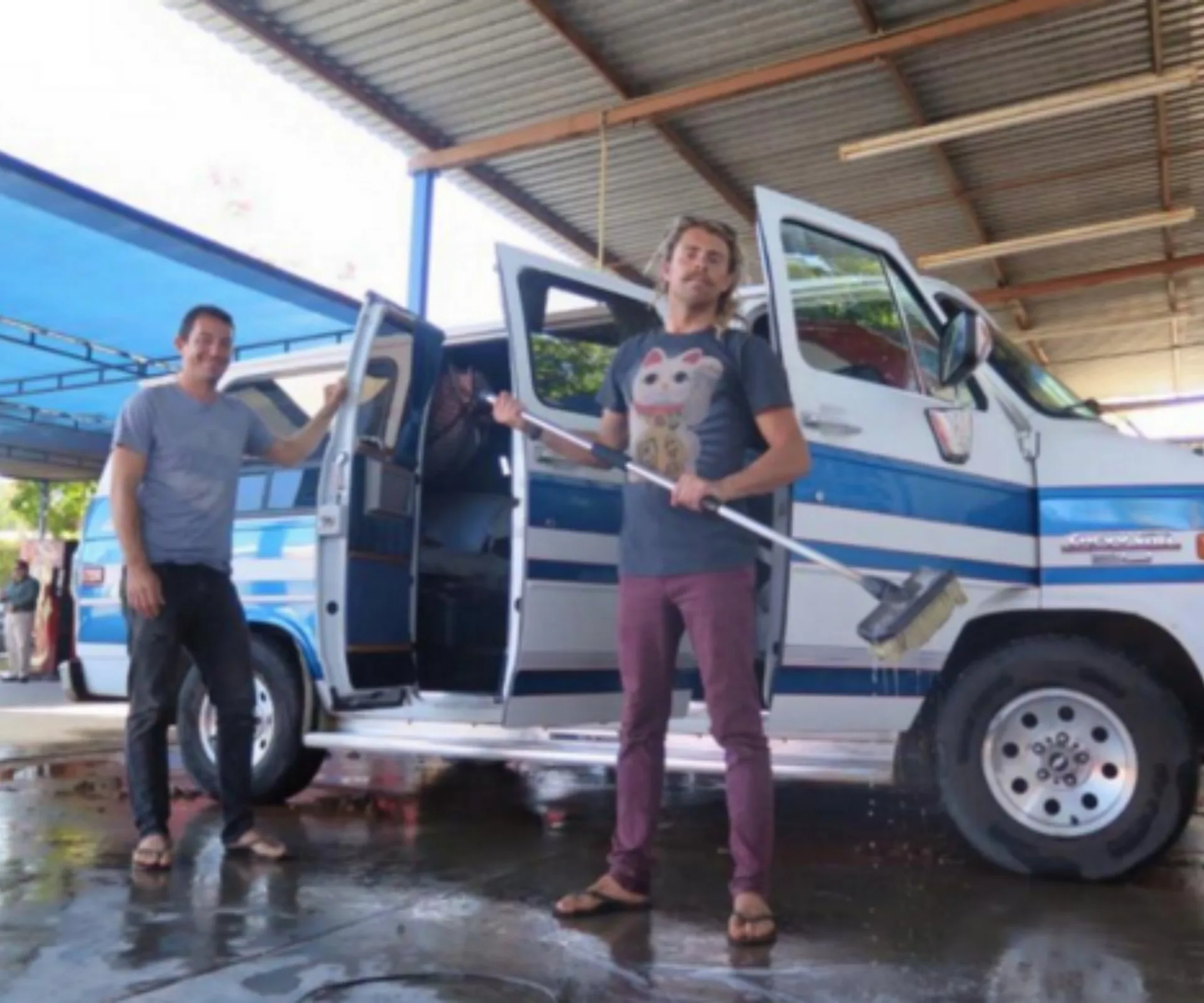 Two people stand by a blue and white van under a roofed area, one holding a broom and smiling.