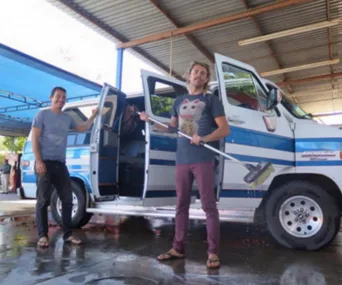Two people stand by a blue and white van under a roofed area, one holding a broom and smiling.