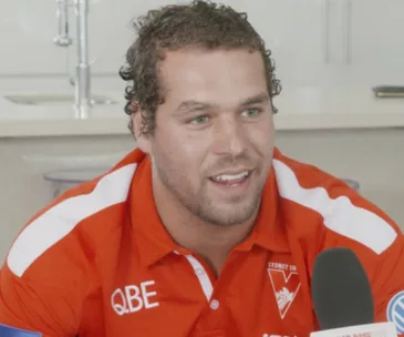 Sydney Swans player speaking to media, wearing a red team shirt with sponsor logos.