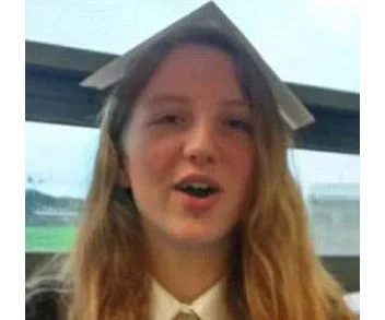 A girl with long hair, wearing a school uniform with a note paper on her head, appears to be speaking indoors.