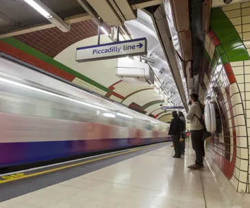 Commuters wait on a London Tube platform as a train speeds by beneath a sign for the Piccadilly line.