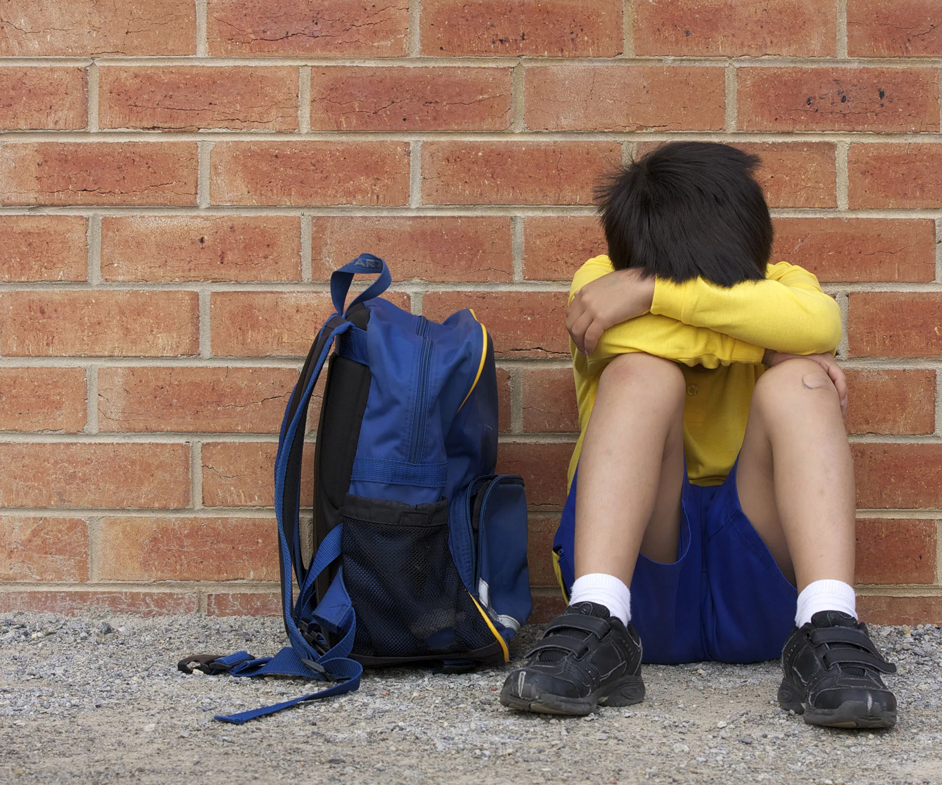 A child in a yellow shirt sits on the ground against a brick wall with a blue backpack beside them, head resting on arms.