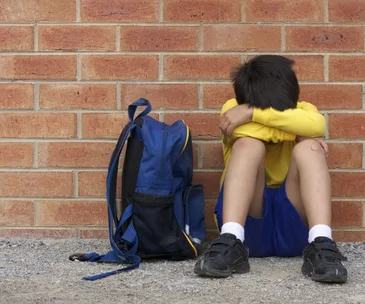 A child in a yellow shirt sits on the ground against a brick wall with a blue backpack beside them, head resting on arms.